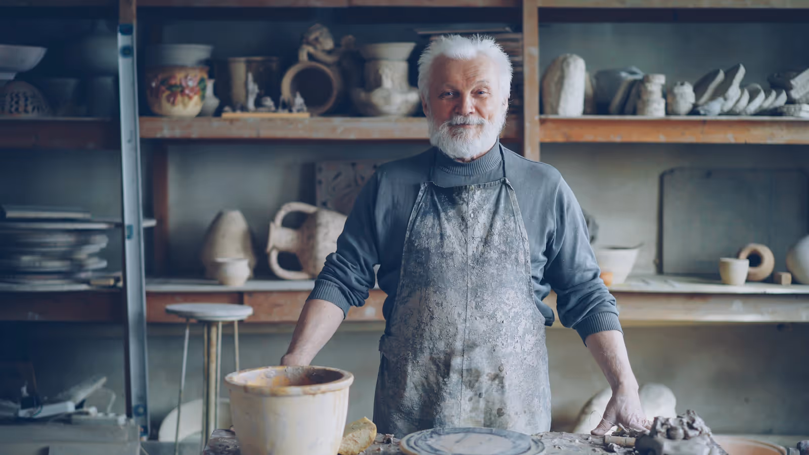 Silver-haired potter and small business owner standing proudly in his workshop, surrounded by handmade pottery